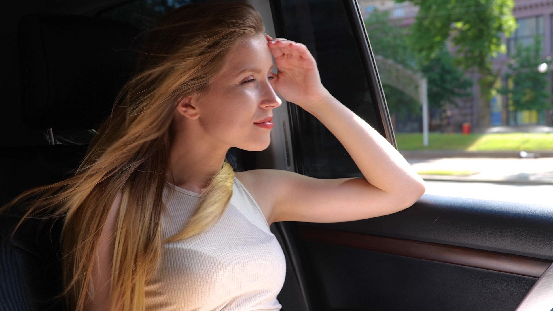 Close up of pretty Caucasian cheerful blonde woman sitting on back seat in a car driving in city. Portrait. Happy beautiful female traveling in auto looking in window on street. Slow motion