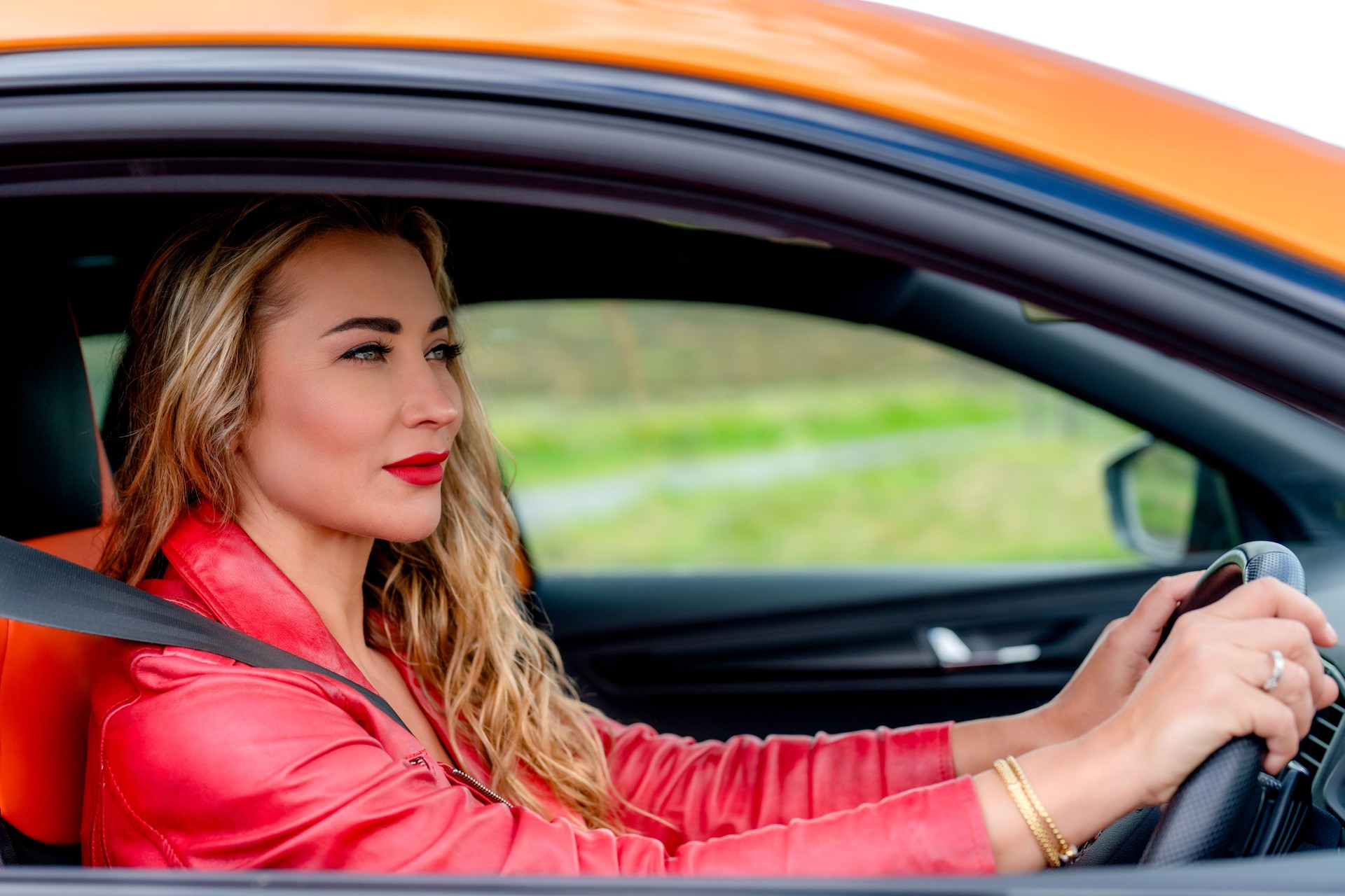 Woman driving an orange car on a scenic road with vibrant greenery in the background during a sunny day